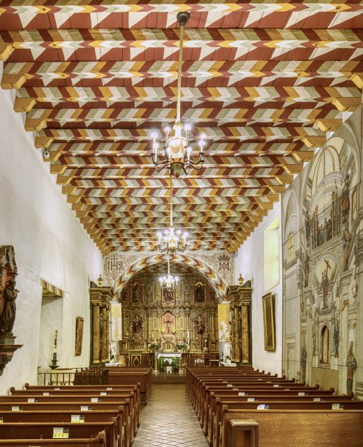 Interior of a church with wooden pews, ornate altar, wall mural, and a ceiling featuring a colorful geometric pattern. Chandeliers hang from the ceiling.