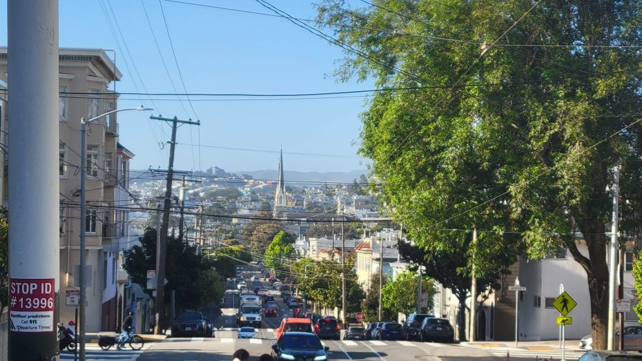 A city street with cars and pedestrians, lined with trees and buildings, slopes downhill toward a distant skyline under a clear blue sky.