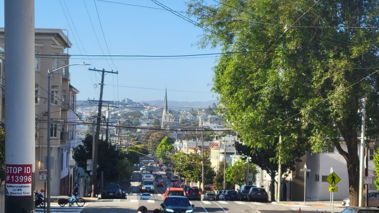 A city street with cars driving and parked along both sides, lined with trees and buildings, with hills and a church spire visible in the background under a clear sky.