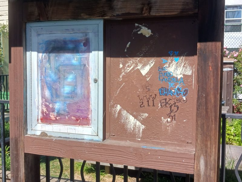 A weathered wooden bulletin board with one painted, closed cabinet and a scratched, graffiti-marked section, situated near a garden and fenced area in sunlight.