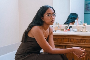 A woman with long dark hair and glasses sits by a bathroom counter with various toiletries, wearing a brown dress and looking at the camera.