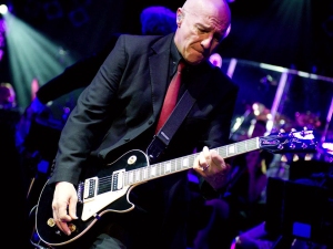 A bald man in a black suit and red tie plays an electric guitar on stage under colorful lights.