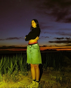 A woman stands outdoors at dusk, wearing a black long-sleeve top, denim skirt, and sneakers, with her arms crossed against a dramatic sunset sky.