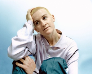 Person with short blonde hair wearing a light-colored shirt and teal pants sits with one hand on their head, looking thoughtfully to the side against a plain background.