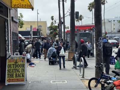 A group of people gather on a city sidewalk near a restaurant sign, with bags and belongings around them; some are walking, others are standing or sitting.