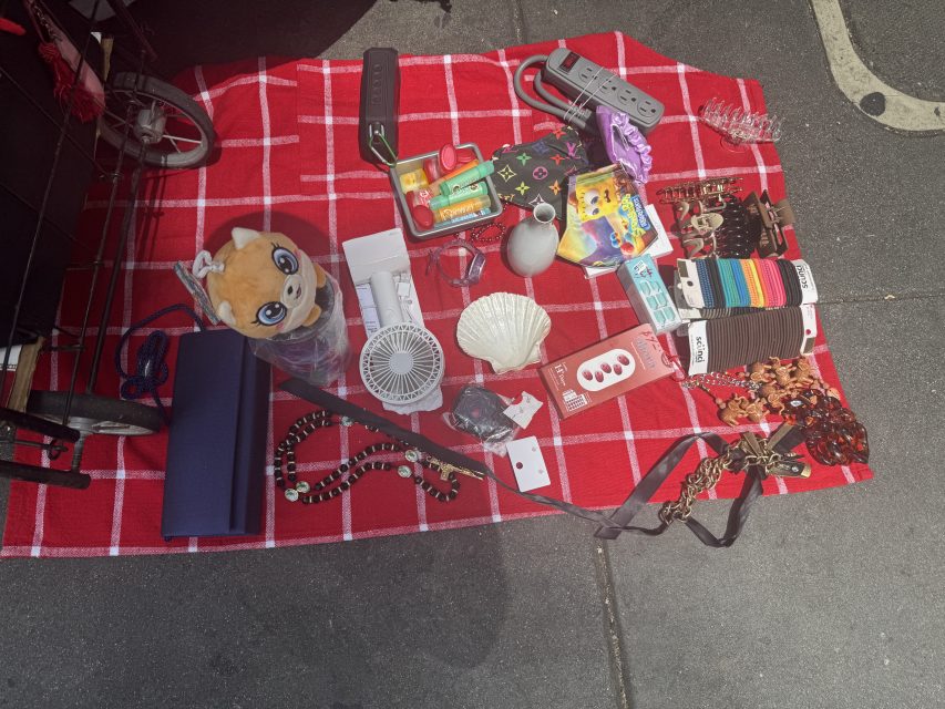 Various items displayed on a red checkered blanket at a sidewalk sale, including plush toy, small fan, belts, jewelry, charger, power strip, and other miscellaneous goods.
