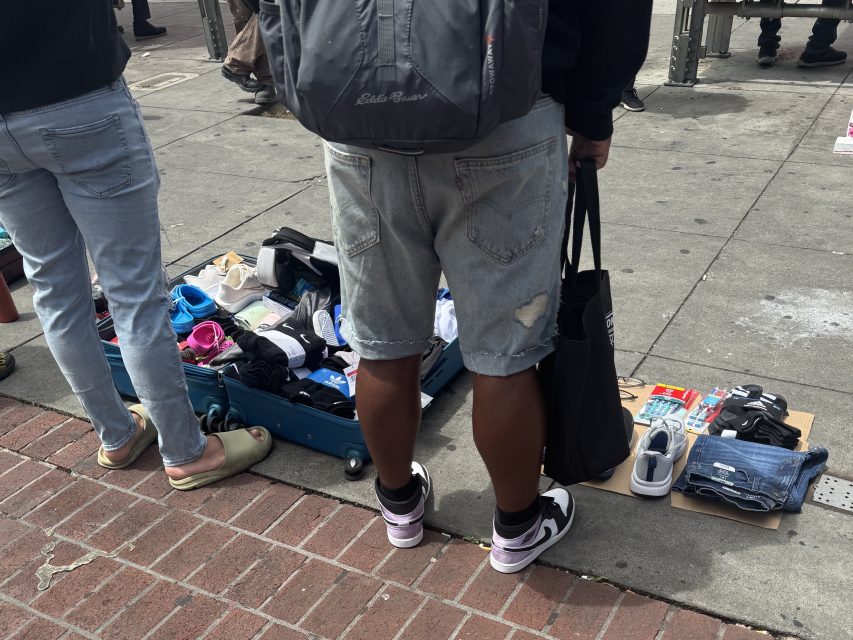People stand on a sidewalk next to a suitcase and cardboard displaying shoes and various items for sale.