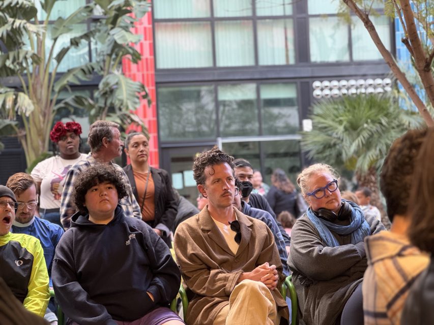 A group of people sit outdoors in chairs, attentively listening. There are trees and a modern building with large windows in the background.