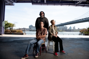 Three people pose under a bridge by the waterfront; two sit on chairs while one stands behind them. City skyline and river are visible in the background.