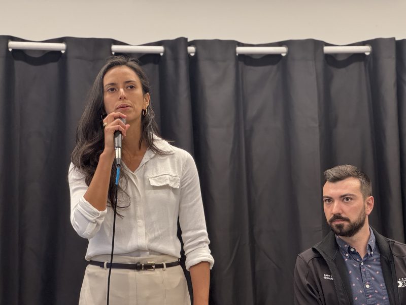 A woman in a white blouse speaks into a microphone while standing beside a seated man with a beard, against a background of black curtains.