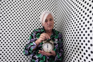An older person with white hair holds a clock in a room with black polka dots on the walls, wearing a floral patterned shirt.