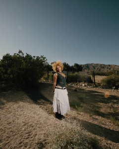 A person with curly blonde hair stands outdoors on sandy ground, wearing a dark sleeveless top and a long white skirt, with shrubs and mountains in the background under a clear sky.