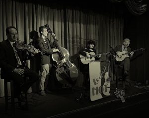 Four musicians perform on stage with a violin, double bass, and two guitars in front of a curtain. A sign reads "Le Jazz Hot." The scene is in black and white.
