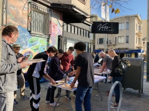 People gather around tables set up on a sidewalk, engaging in activities or filling out forms in front of a building with a mural and a sign that reads "Vita Wellness.