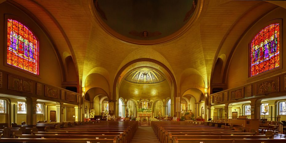 Interior of a church with wooden pews, arched ceiling, stained glass windows, and an ornate altar illuminated by warm lighting.