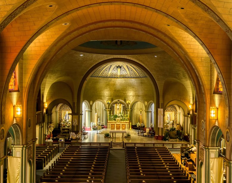 Interior of a large, ornate church with wooden pews, arched ceilings, stained glass windows, and an illuminated altar at the front.