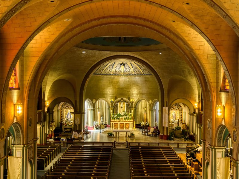 Interior of a large, ornate church with wooden pews, arched ceilings, stained glass windows, and an illuminated altar at the front.
