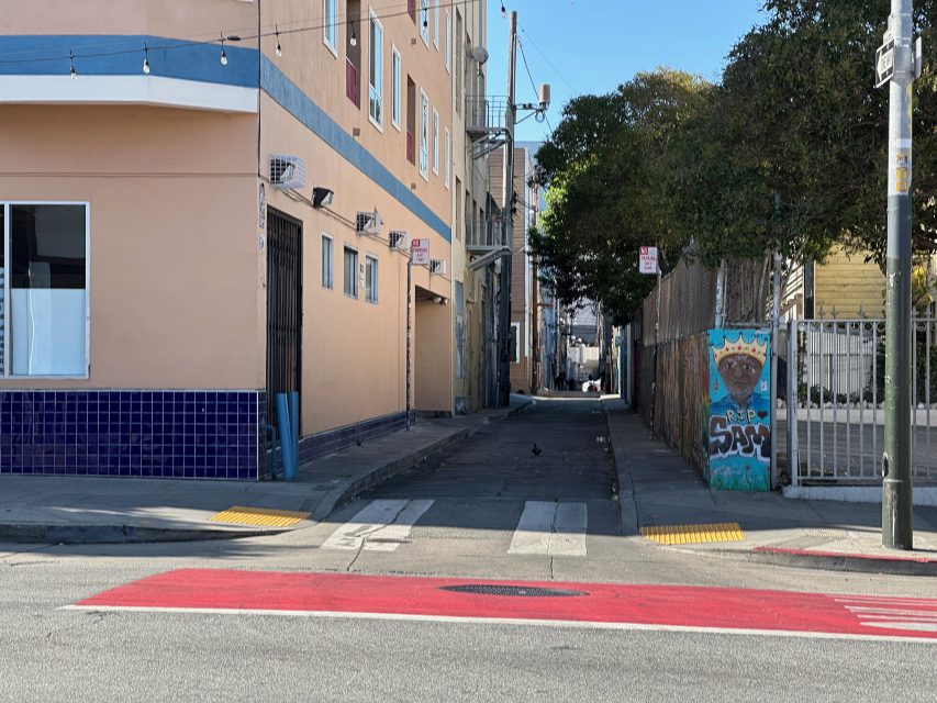 A narrow alleyway between buildings, with a painted mural on the right wall and a small crosswalk in the foreground under clear daylight.