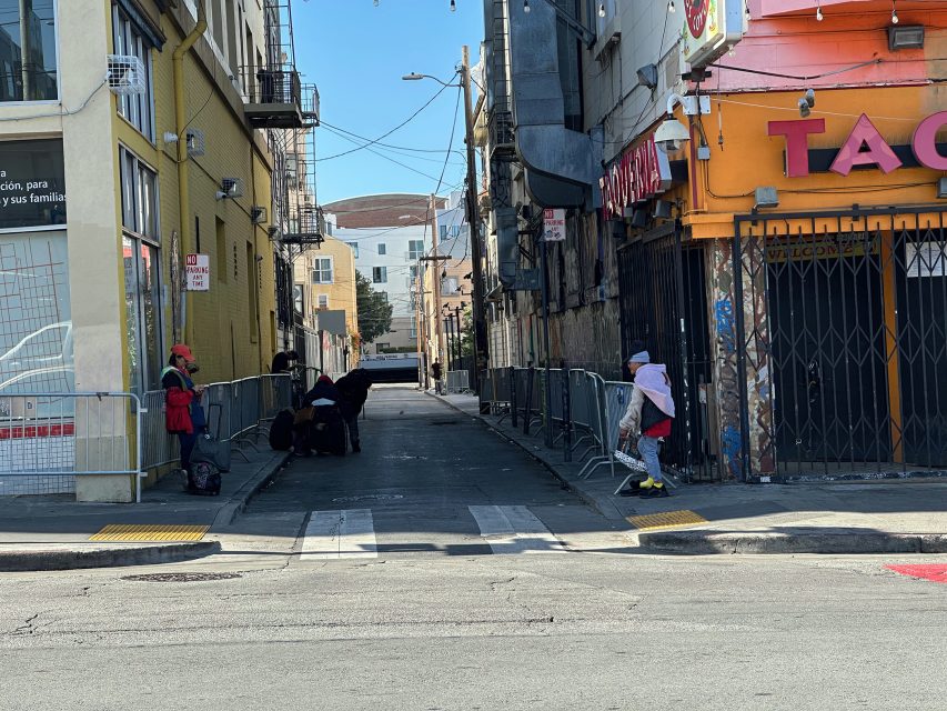 A fenced alleyway with several people gathered near the entrance; one person walks past, and nearby buildings have closed gates and signs.