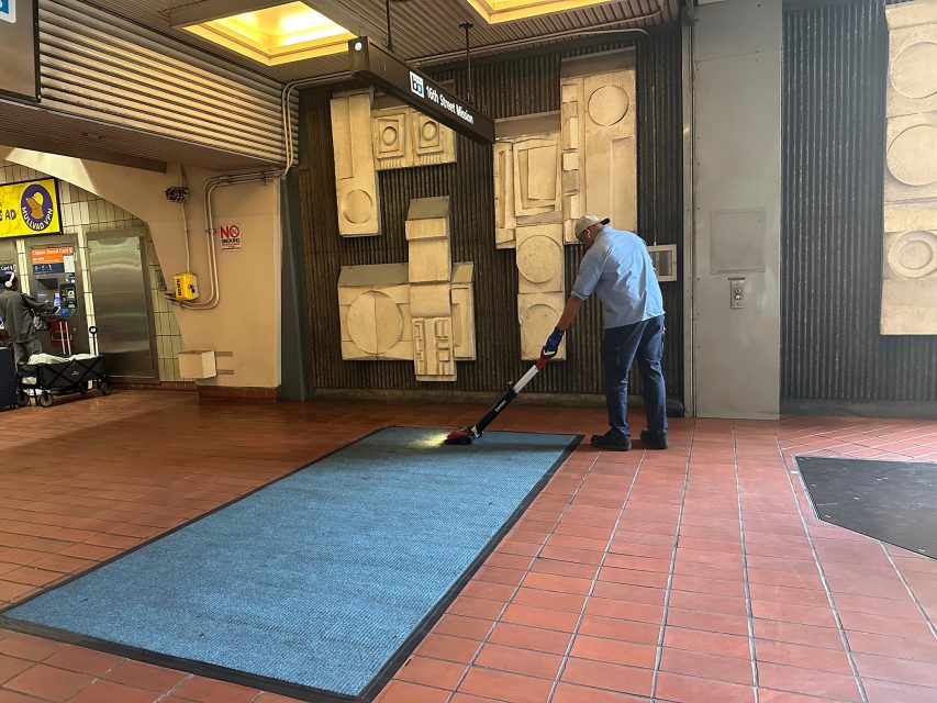 A person in a blue shirt and hat is vacuuming a blue rug in a tiled indoor public area with abstract wall art.