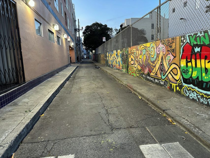 Narrow urban alley with colorful graffiti on a fence, buildings on the left, and a cracked asphalt road under a dim evening sky.