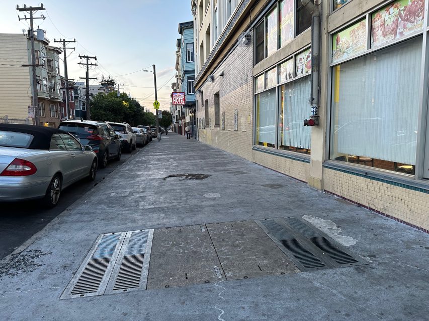 A city sidewalk lined with parked cars next to buildings, including one with large windows and posters, under a clear sky.