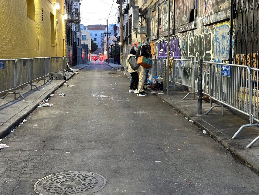 Two people stand near a graffitied wall in an empty, litter-strewn alley lined with metal barricades and buildings, as evening approaches.