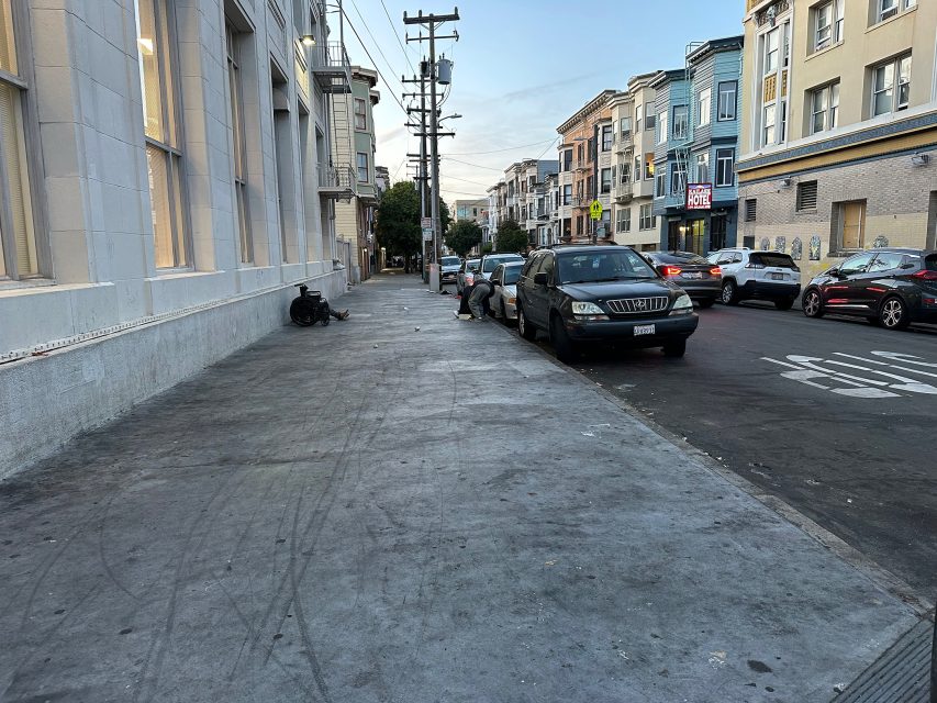 A wide, empty sidewalk runs alongside parked cars and multi-story buildings on a city street in the early evening.
