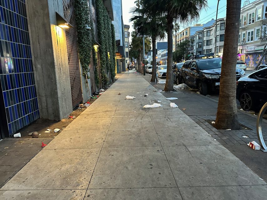 A city sidewalk lined with trees, scattered litter and debris on the ground, with parked cars and buildings on both sides.