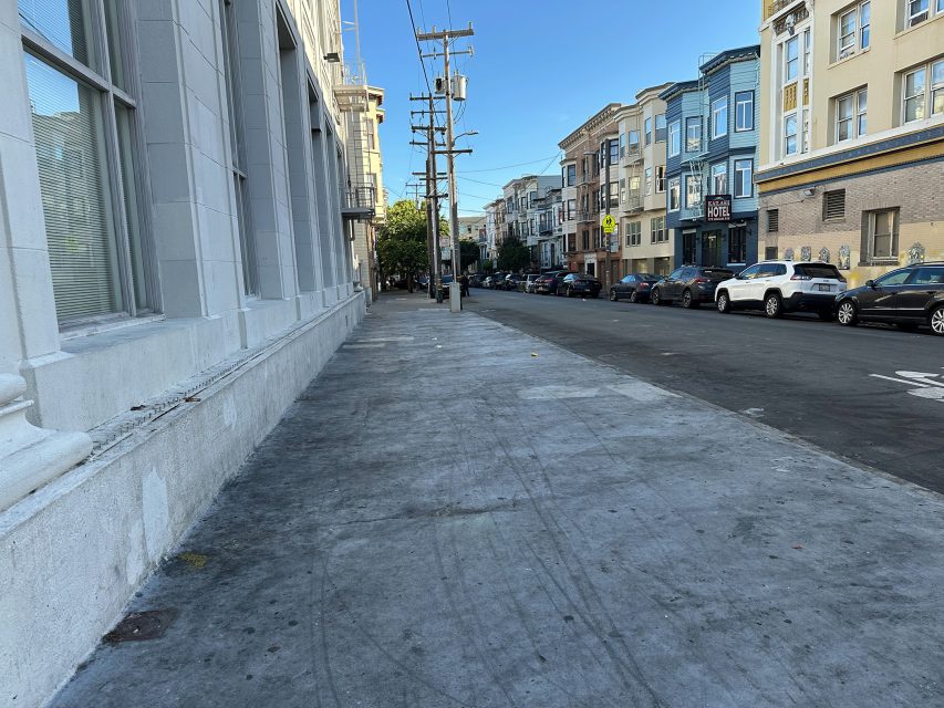 A quiet city street with parked cars, multi-story buildings, utility poles, and a wide, empty sidewalk under a clear blue sky.