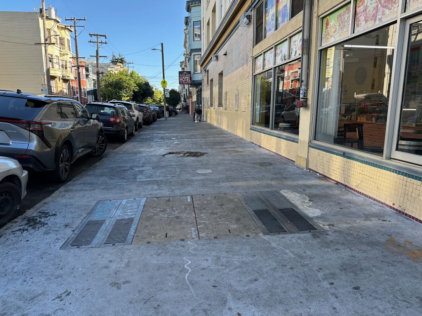 A sidewalk next to parked cars and a building with large windows; metal grates are embedded in the sidewalk.