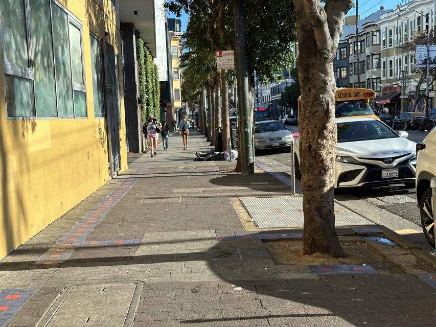 A city sidewalk with pedestrians, parked cars, a school bus, and a person lying on the ground in the shade of a tree on a sunny day.