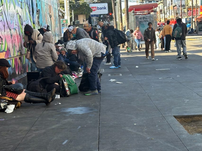 A group of people gather and sit on a city sidewalk near a graffiti-covered wall; some are seated with bags and belongings, while others stand or walk by.