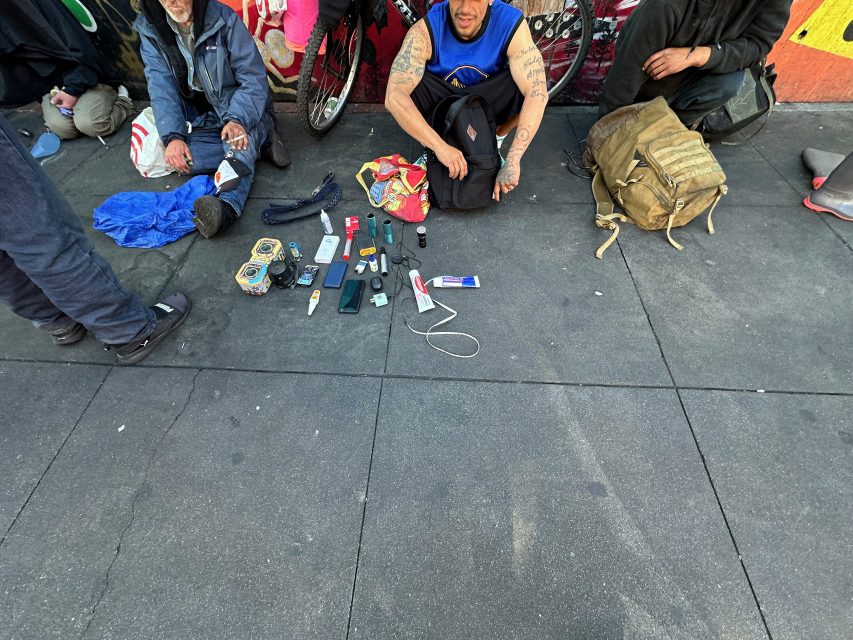 Several people sit on a sidewalk with various personal items, electronics, and toiletries arranged on the ground in front of them.