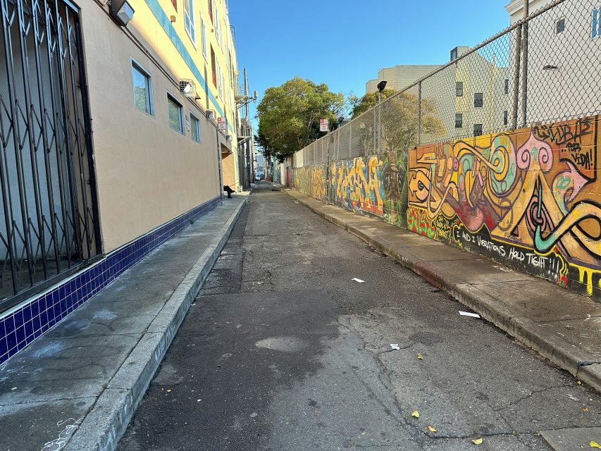 A narrow urban alley with graffiti-covered walls, a metal gate on the left, and a chain-link fence on the right, under a clear blue sky.