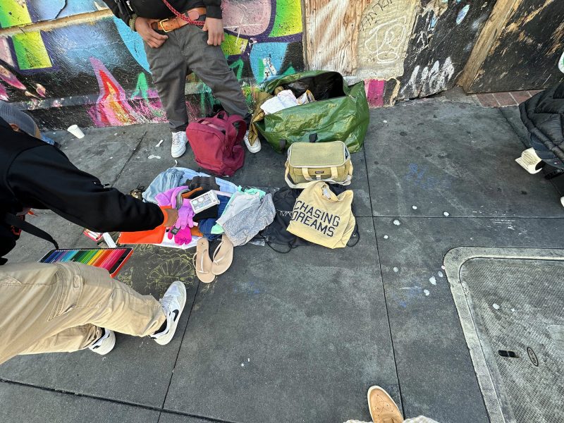 Two people stand on a city sidewalk next to bags, scattered clothes, art supplies, and a tote bag labeled "Chasing Dreams" near a colorful graffiti-covered wall.