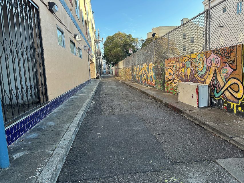 Narrow urban alley with graffiti-covered wall on the right, chain-link fence, and buildings on the left; clear sky visible above.