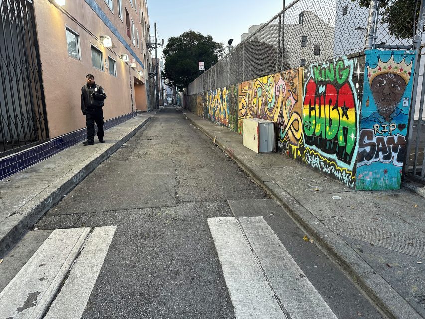 A man in dark clothing stands on the left side of an empty urban alley with colorful graffiti murals on a fence to the right.