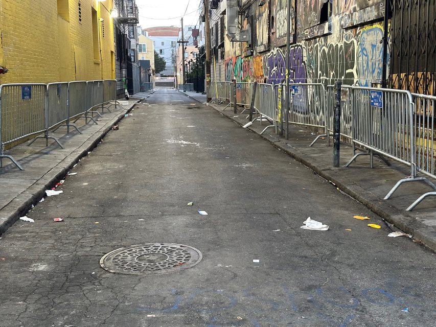 Narrow urban alleyway with metal barricades on both sides, graffiti on walls, litter scattered on the ground, and buildings in the background.
