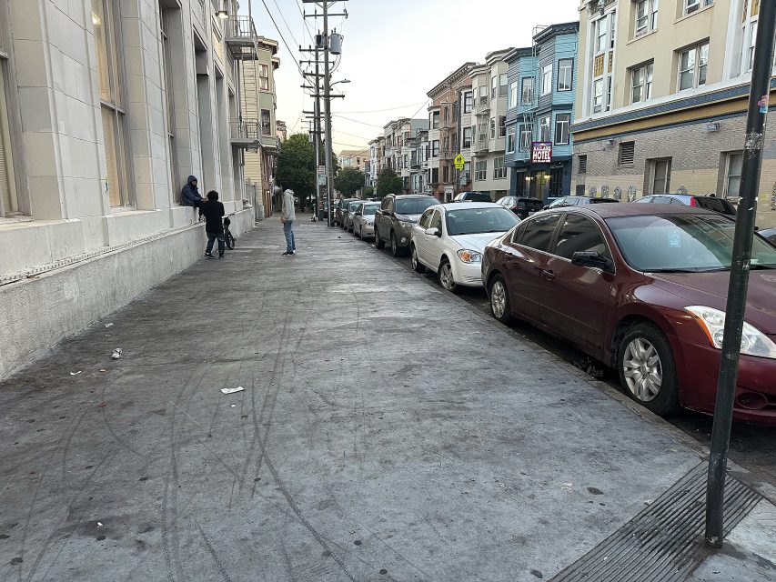 A city street with parked cars on the right, several people near a building on the left, and multi-story buildings lining both sides.