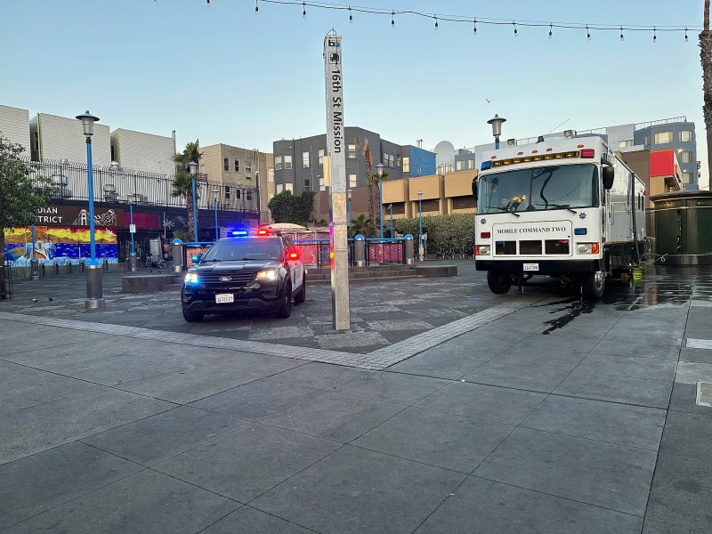 A police car with flashing lights and a mobile command vehicle are parked in a city plaza near buildings and street art.