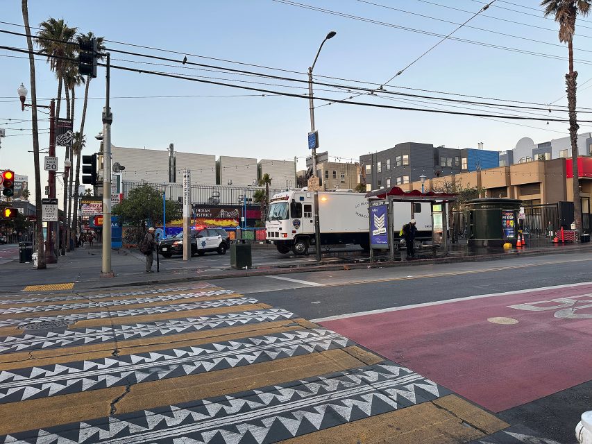 A city street intersection with a patterned crosswalk, a street cleaning vehicle, and people waiting at a bus shelter in an urban area with colorful buildings.