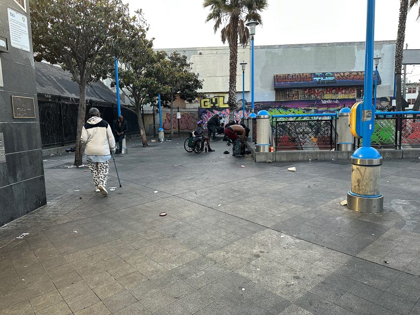 Urban street scene with several people, some sitting or standing near graffiti-covered walls and trash cans, and one person walking with a cane on a paved area.