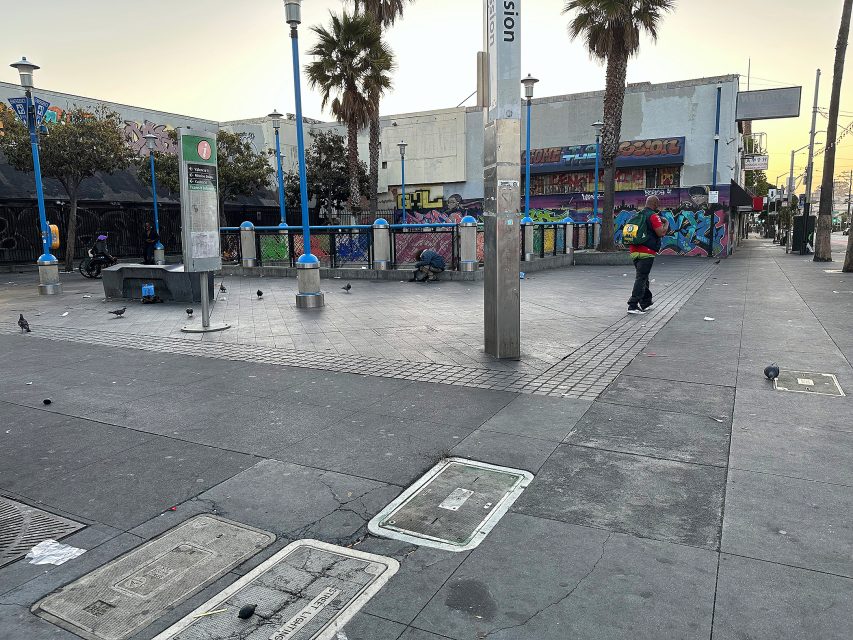 Urban plaza with scattered pigeons, a few people, palm trees, street art on building walls, and empty benches on a mostly deserted city street.