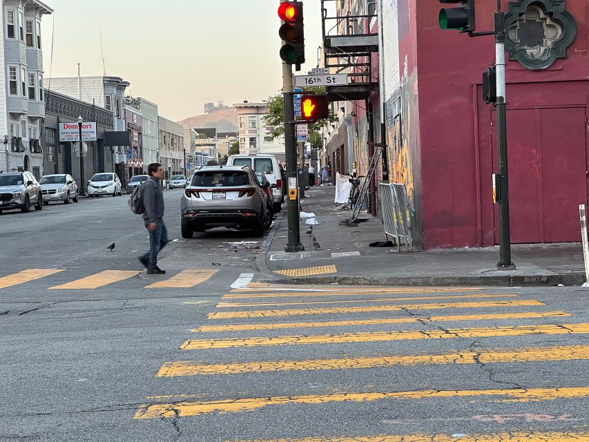 A man with a backpack waits at a crosswalk on 16th Street; the traffic light is red and cars are parked along the street.