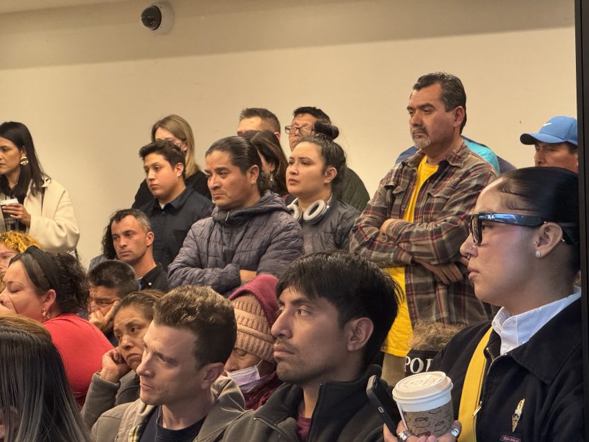A diverse group of people stands and sits closely together in a crowded indoor room, some with serious expressions, others holding drinks and wearing jackets.