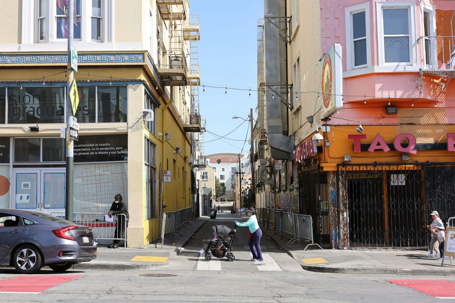 A person pushes a stroller across a city street in front of an alleyway, with shops and buildings on either side and a parked car on the left.