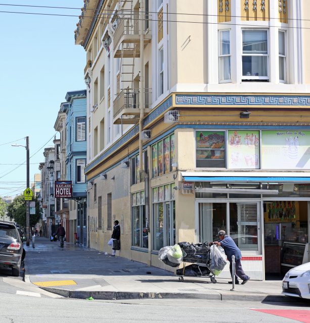 A person pushes a shopping cart filled with bags past a corner store and colorful buildings in an urban neighborhood.