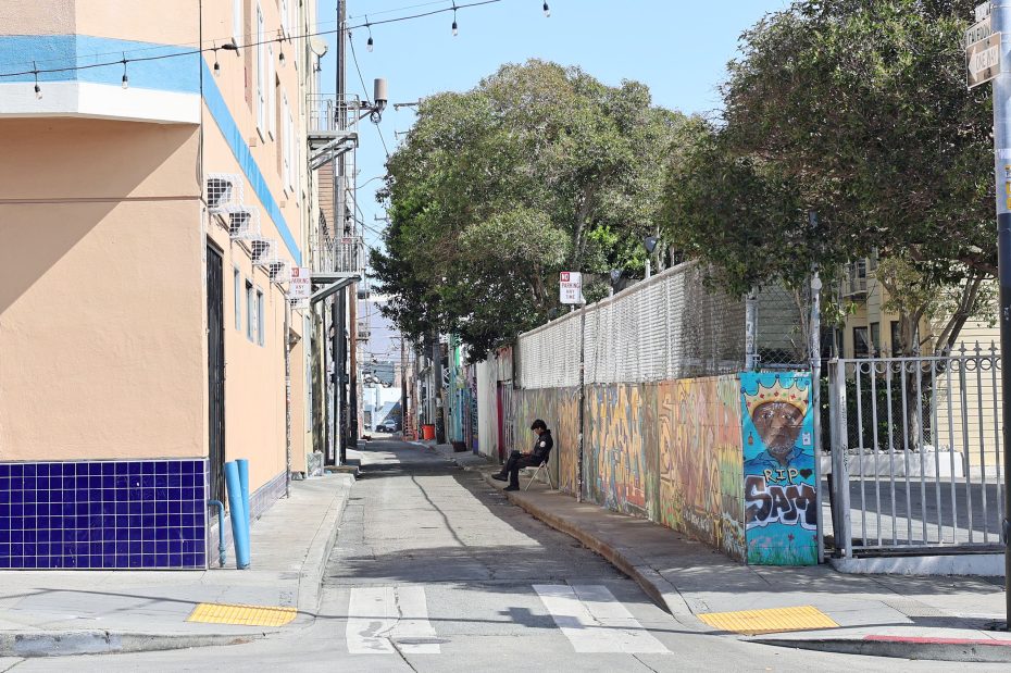 A narrow urban alley with murals on the walls, trees in the background, and a person sitting alone on the sidewalk.