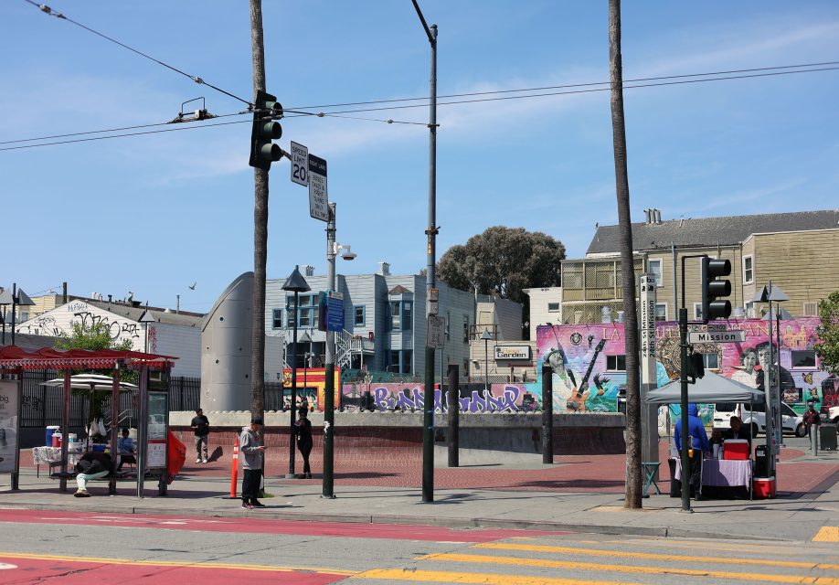 A city street scene shows a bus stop, people walking, colorful murals, traffic lights, and buildings under a clear sky.